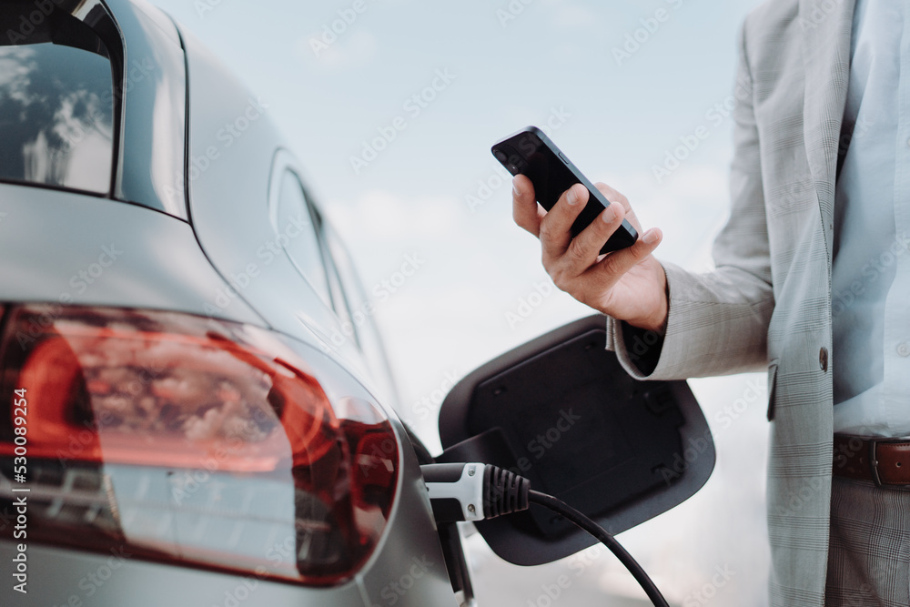 Man holding smartphone while charging car at electric vehicle charging station, closeup. - obrazy, fototapety, plakaty 
