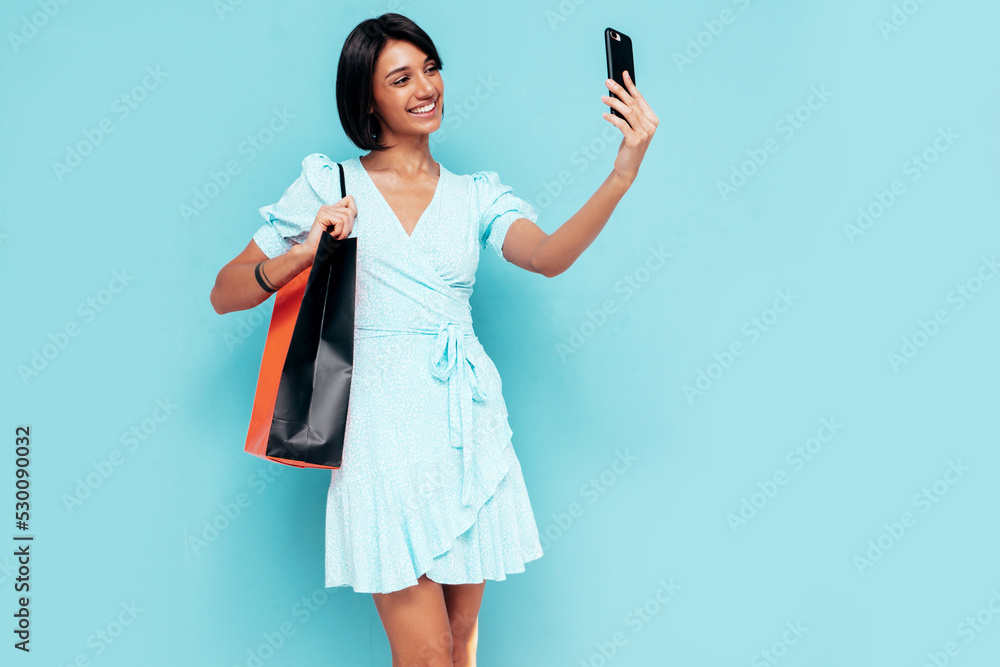 Young beautiful smiling female in trendy summer dress. Carefree woman posing near blue wall in studio. Positive model holding shopping bag. Cheerful and happy. Taking selfie Young beautiful smiling female in trendy summer dress. Carefree woman posing near blue wall in studio. Positive model holding shopping bag. Cheerful and happy. Taking selfie