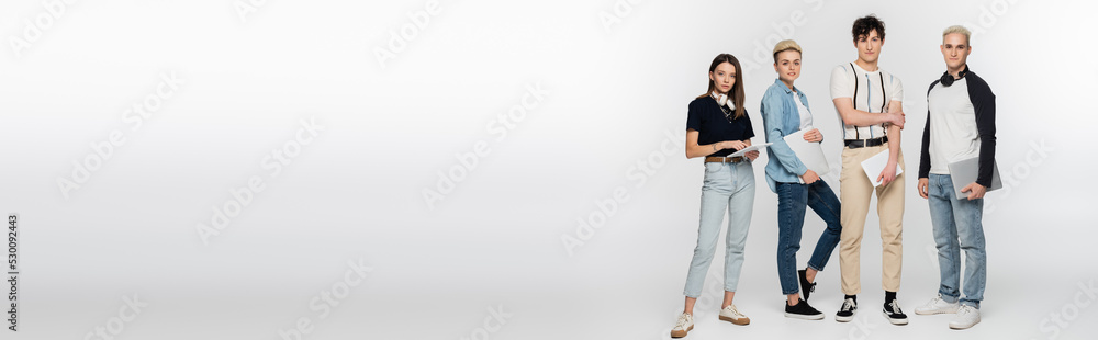 full length of young people with digital tablets and laptops looking at camera on grey background, banner. full length of young people with digital tablets and laptops looking at camera on grey background, banner.