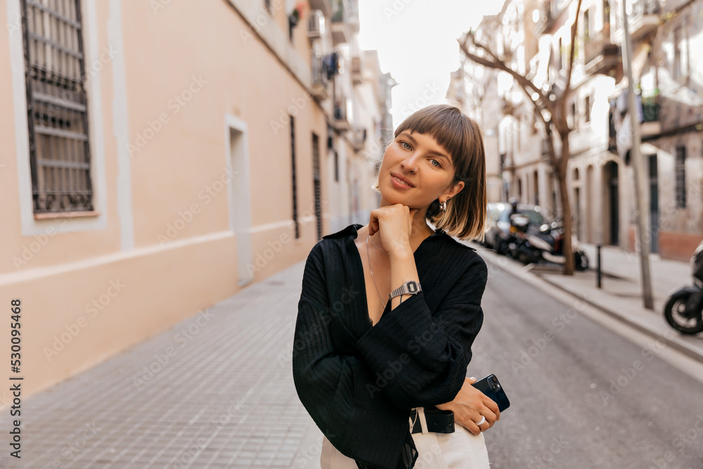 Romantic lovely girl with short hair wearing black shirt walking on the city in summer sunny evening. Brown-haired girl with smartphone wears black shirt. Happy weekend concept.