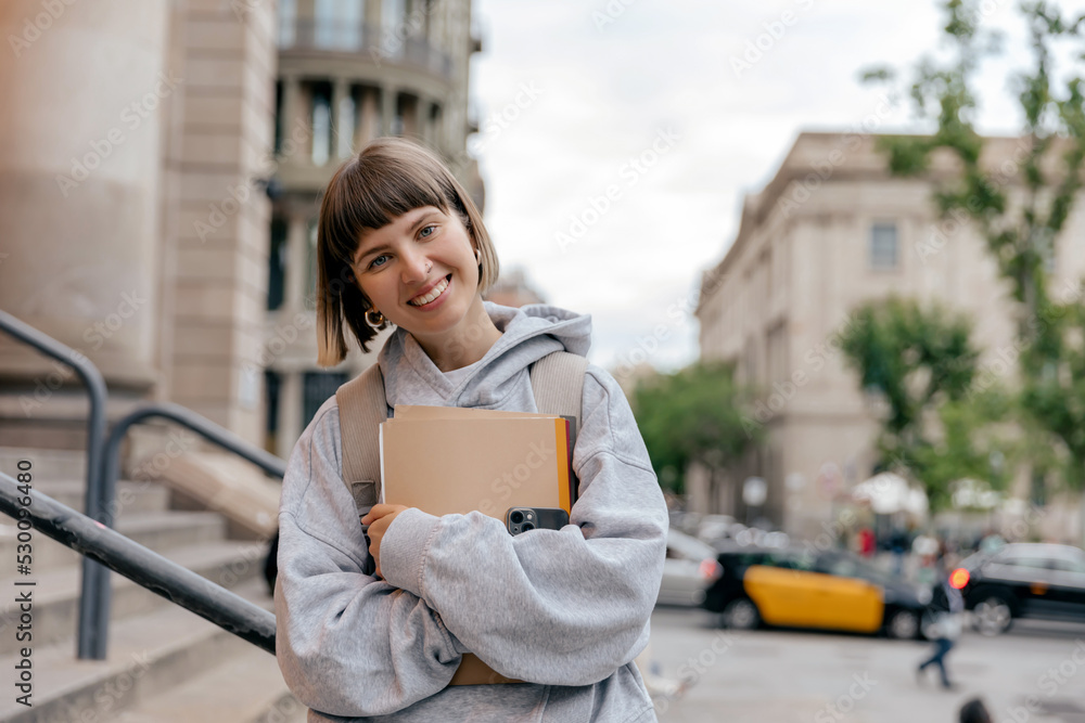 Lovely smiling adorable lady with wonderful smile posing at camera with notes and backpack on city background