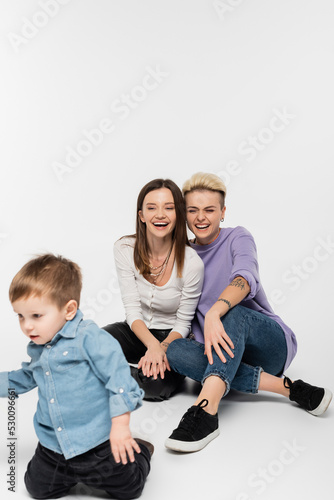 cheerful lesbian couple laughing near toddler son on grey background.