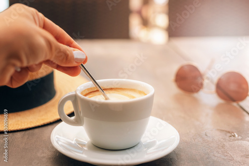 Cappuccino on the table in cafe. Women's hands stirs the sugar in coffee. Morning sunlight falls on table. Breakfast concept. Selective focus