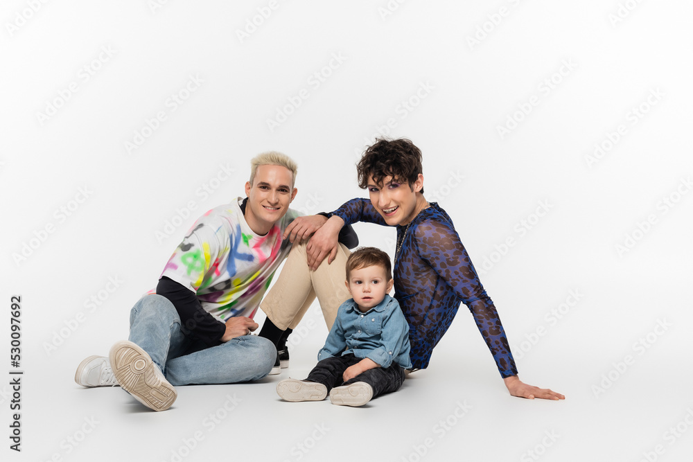stylish gay partners sitting near toddler son on grey background. stylish gay partners sitting near toddler son on grey background.