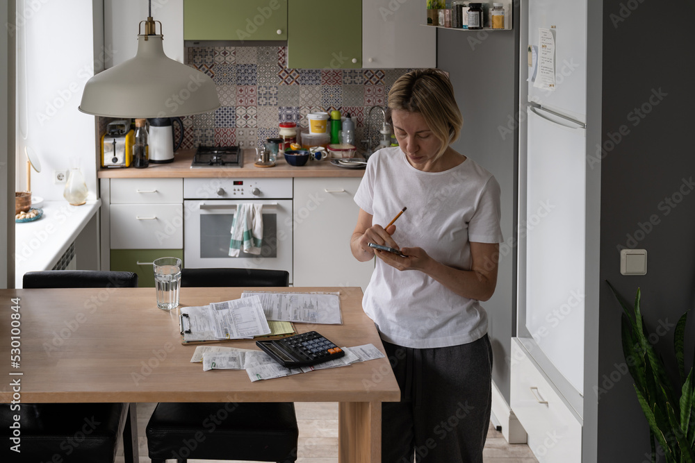 Busy woman with bunch of unpaid bills on table tries to solve problems with obligations standing in kitchen. Middle aged female wants to pay bills searching contacts of hotline on Internet on phone Busy woman with bunch of unpaid bills on table tries to solve problems with obligations standing in kitchen. Middle aged female wants to pay bills searching contacts of hotline on Internet on phone