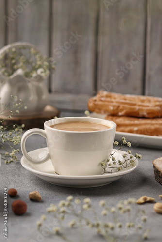 A white cup of coffee with milk stands on a gray background, behind a plate with eclairs