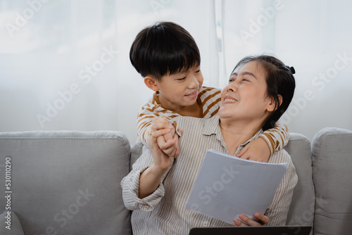 Asian single mom sitting on vacation working, His naughty son stood behind the sofa, and covered his mother's eyes with his hand, he tried to play with his mother, Mother turned around and smiled.