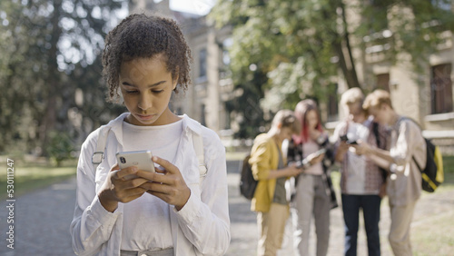 Sad African American girl suffering from social media bullying from group of school children