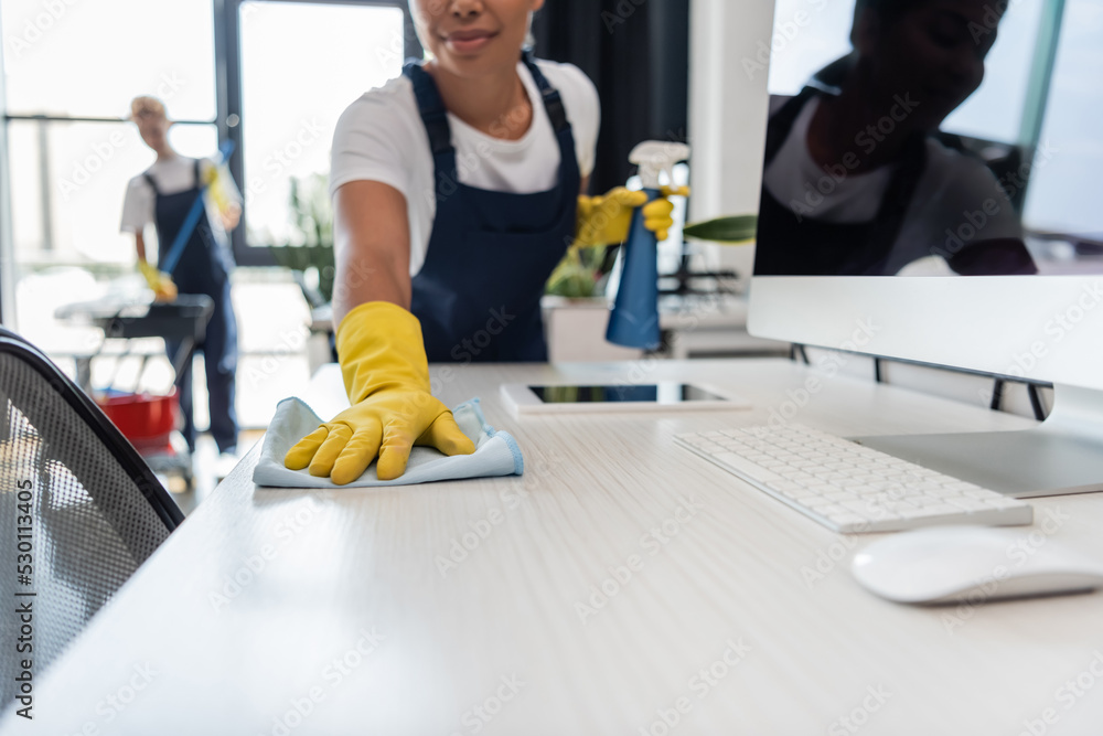 cropped view of bi-racial woman in rubber gloves cleaning office desk near gadgets and colleague on blurred background. cropped view of bi-racial woman in rubber gloves cleaning office desk near gadgets and colleague on blurred background.