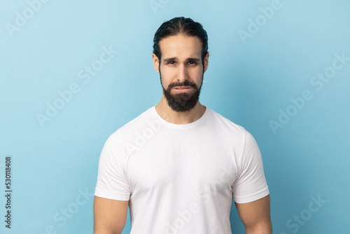 Portrait of man with beard wearing white T-shirt standing and looking at camera with dissatisfied sadness face, expressing sorrow, having bad mood. Indoor studio shot isolated on blue background.