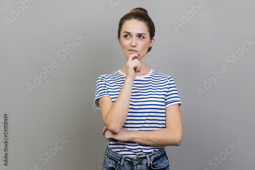 Photos Portrait of pensive woman wearing striped T-shirt standing holding chin, thinking about important question, having thoughtful facial expression