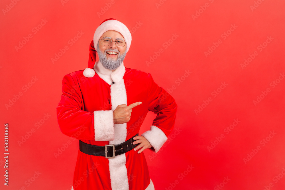 Satisfied elderly man with gray beard wearing santa claus costume standing with hand on hips, pointing index finger aside, presenting copy space. Indoor studio shot isolated on red background. Satisfied elderly man with gray beard wearing santa claus costume standing with hand on hips, pointing index finger aside, presenting copy space. Indoor studio shot isolated on red background.