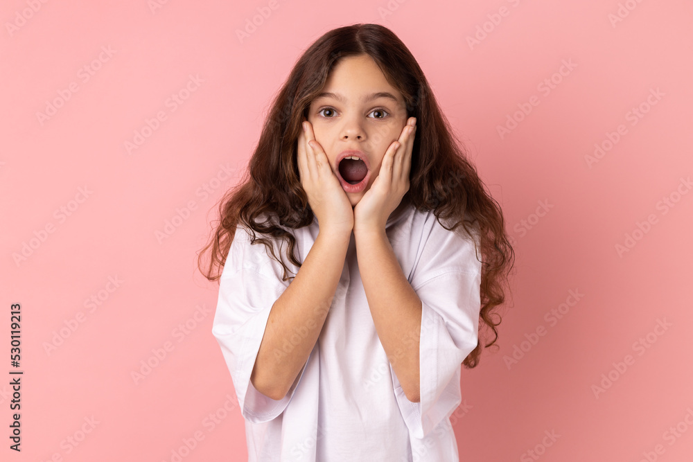 Portrait of amazed little girl wearing white T-shirt standing with hands on cheeks, looking at camera with big eye, saying wow. Indoor studio shot isolated on pink background. Portrait of amazed little girl wearing white T-shirt standing with hands on cheeks, looking at camera with big eye, saying wow. Indoor studio shot isolated on pink background.