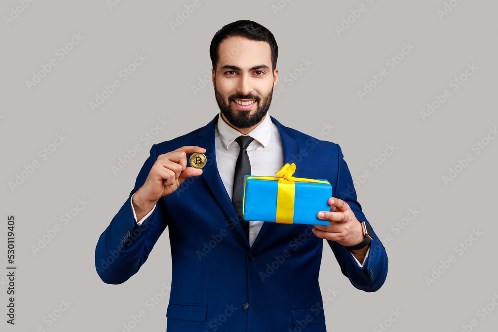 Smiling positive bearded businessman standing holding present box and golden bitcoin, crypto currency as a gift, wearing official style suit. Indoor studio shot isolated on gray background. Smiling positive bearded businessman standing holding present box and golden bitcoin, crypto currency as a gift, wearing official style suit. Indoor studio shot isolated on gray background.