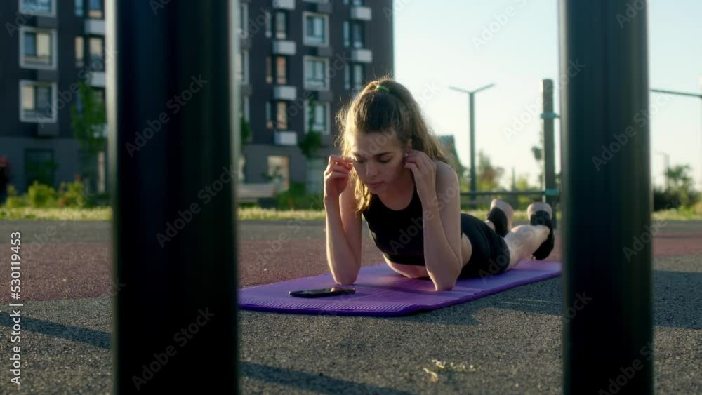 pretty slender young woman puts headphones in her ears after or before exercising while lying on gymnastic mat early in the morning outdoors on sports field in yard of her residential complex. pretty slender young woman puts headphones in her ears after or before exercising while lying on gymnastic mat early in the morning outdoors on sports field in yard of her residential complex.