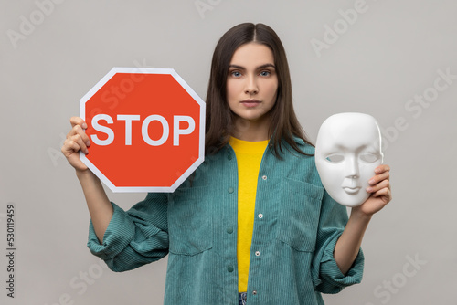 Portrait of serious dark haired woman holding red stop sign and white mask, looking at camera with strict expression, wearing casual style jacket. Indoor studio shot isolated on gray background. photo