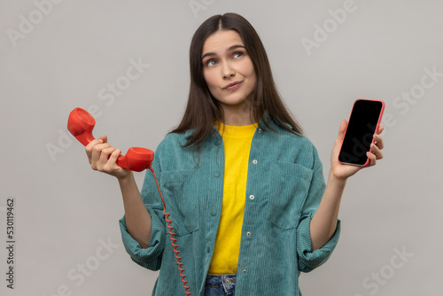 Portrait of confused puzzled woman standing retro telephone and mobile phone, thinking what to use better, wearing casual style jacket. Indoor studio shot isolated on gray background. photo