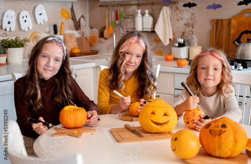 Three cute little sisters drawing and carving spooky faces on pumpkins in light white kitchen at home. Halloween party preparation.