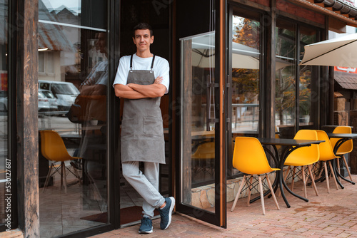 barista or waiter cafe or coffee shop owner against entrance, gesture inviting you to visit, smiling guy in apron standing outdoors being proud of his small local business