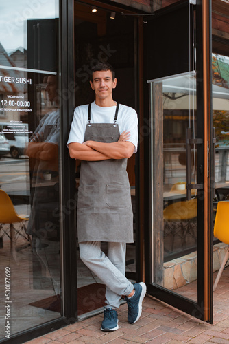 barista or waiter cafe or coffee shop owner against entrance, gesture inviting you to visit, smiling guy in apron standing outdoors being proud of his small local business © shangarey