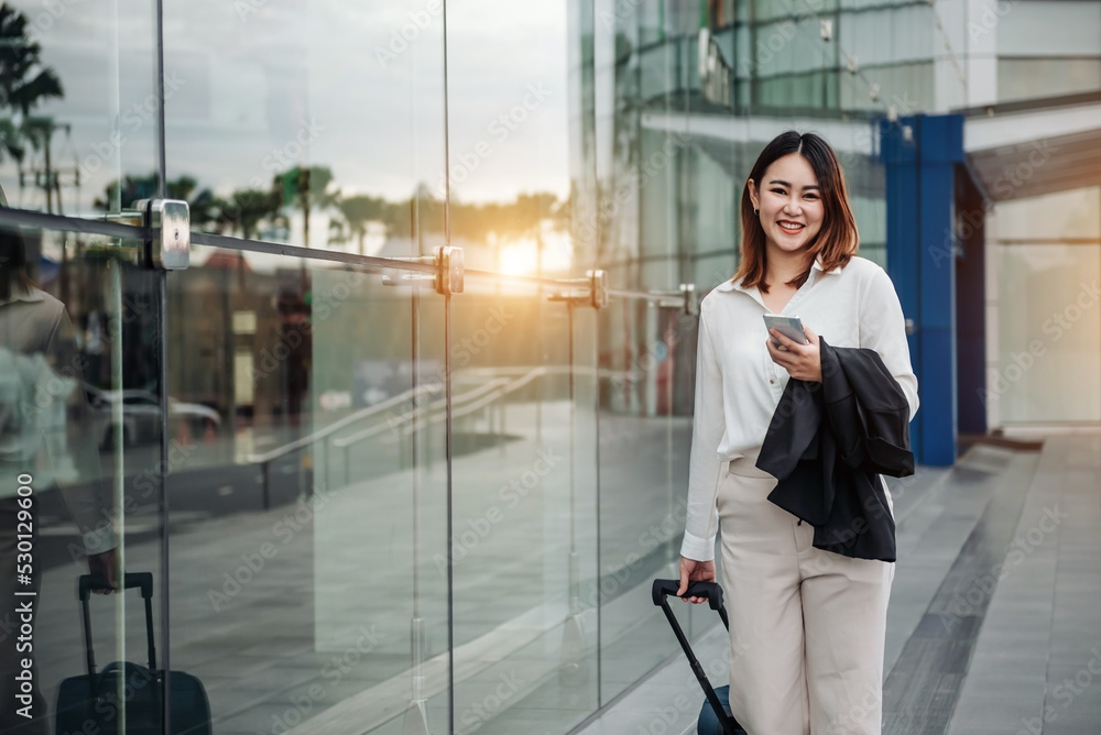 Fototapeta premium Asian woman on smart phone at gate waiting in terminal. Air travel concept with young casual business woman sitting with talking on the smartphone, carry-on hand luggage trolley.