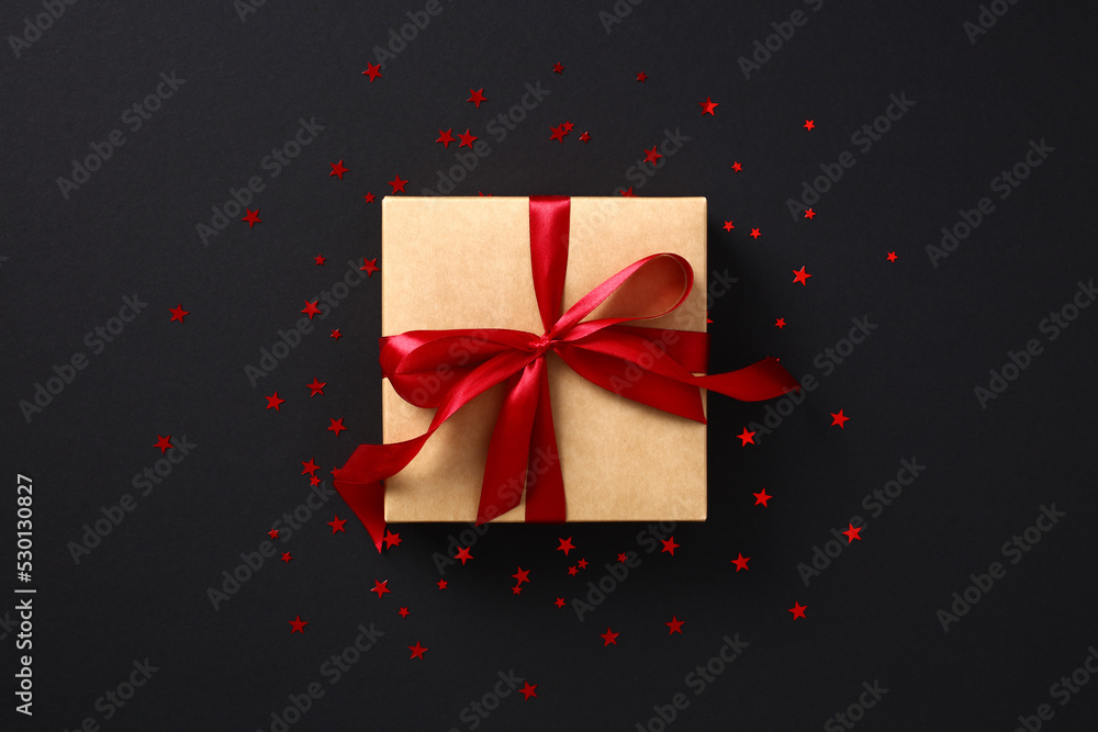 Luxury craft paper gift box with red ribbon bow with confetti stars on black background. Black Friday sale concept. Luxury craft paper gift box with red ribbon bow with confetti stars on black background. Black Friday sale concept.