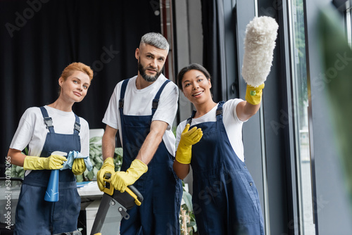 cheerful bi-racial woman pointing with dust brush near colleagues with cleaning supplies.