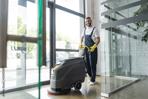 full length of professional cleaner with electrical floor scrubber machine smiling at camera.