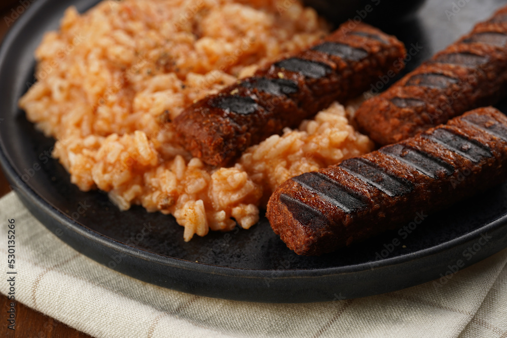 Traditional south european skinless sausages cevapcici made of ground meat and spices on black plate on dark wooden board, vegan substitute made of soy protein, with rice and sour cream Traditional south european skinless sausages cevapcici made of ground meat and spices on black plate on dark wooden board, vegan substitute made of soy protein, with rice and sour cream