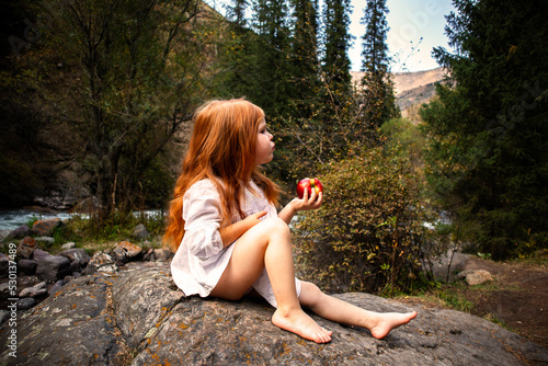 a little girl with long red hair in a white dress is sitting near the river and eating an apple