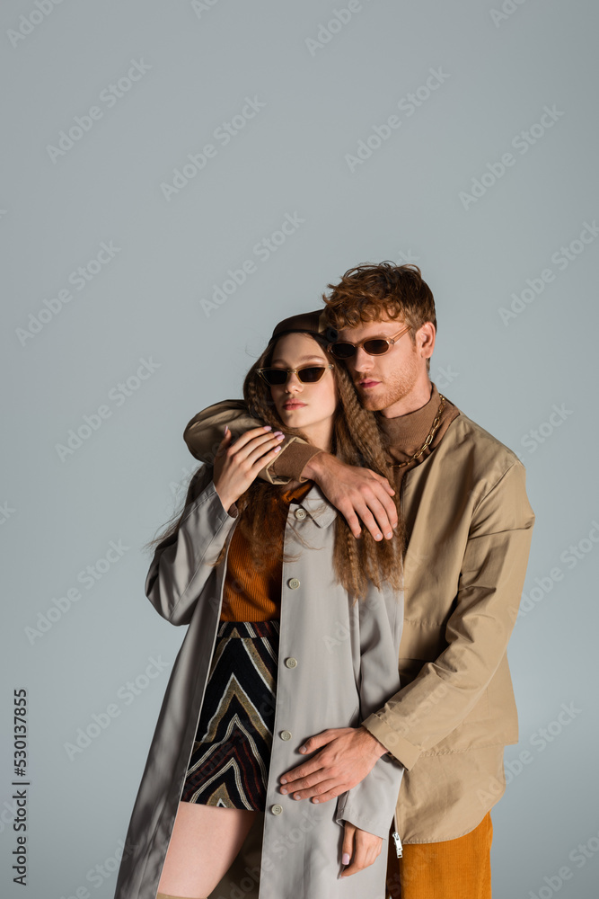young redhead man in sunglasses hugging girlfriend in headscarf isolated on grey. young redhead man in sunglasses hugging girlfriend in headscarf isolated on grey.