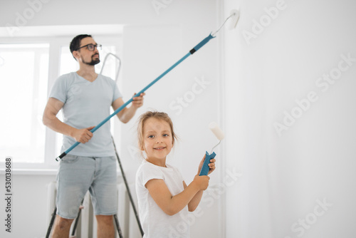 Smiling 4-year-old girl holds a paint roller in her hands. The child helps her father, who is standing behind her, near the wall, in the repair.