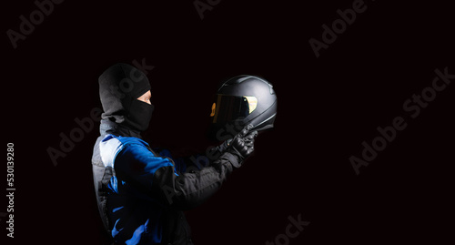 male motorcyclist in a motorcycle outfit and a balaclava puts on a helmet on a dark background. The concept of protective equipment for a biker. © velimir