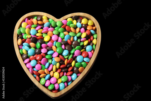 735 / 5.000 Resultados de tradução Chocolate candies covered in colored sugar in a wooden heart-shaped bowl isolated over a black background. Top view.