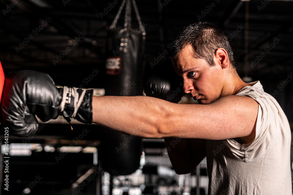High quality photography. Caucasian man in profile with arm outstretched throwing a punch while boxing with a punching bag. Short-haired man in a gray tank top training.