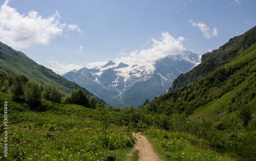 Beautiful landscape - the slopes of high mountains and the green Alpine valley of Terskol with a walking path in the Elbrus region in Kabardino-Balkaria on a clear summer sunny day