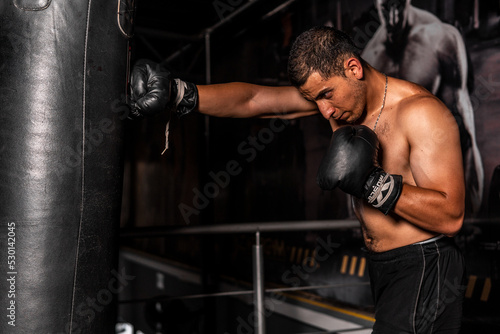 High quality photography. Shirtless Latino man pounding a punching bag. A Latin man training box inside a dark gym.