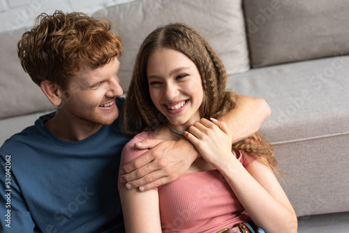 redhead and happy young man hugging and looking at cheerful girlfriend in living room.
