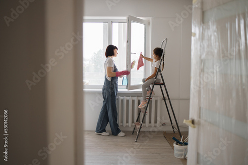 Mother and daughter child in rubber gloves with detergent and rag washing windows together. Girl helping woman do house cleaning after repair.