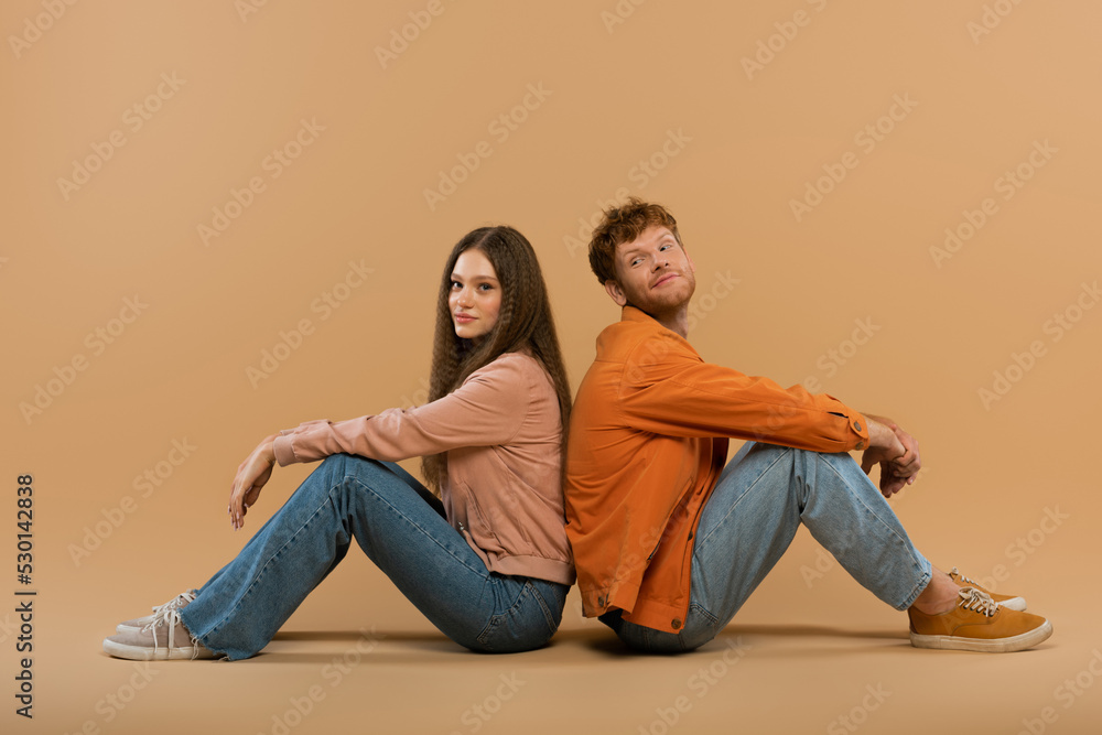 full length of smiling young man with red hair sitting back to back with girlfriend on beige. full length of smiling young man with red hair sitting back to back with girlfriend on beige.