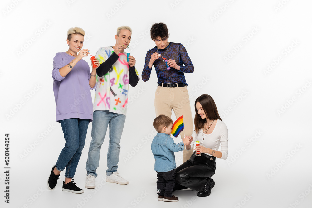 young lgbtq community friends blowing soap bubbles near toddler boy on grey background. young lgbtq community friends blowing soap bubbles near toddler boy on grey background.