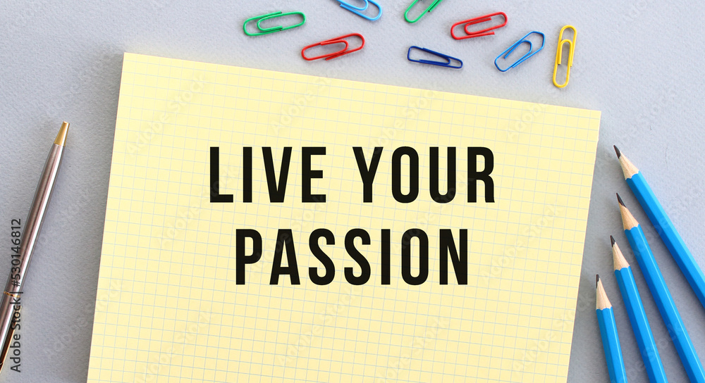 LIVE YOUR PASSION text in notebook on gray background next to pencils, pen and paper clips.