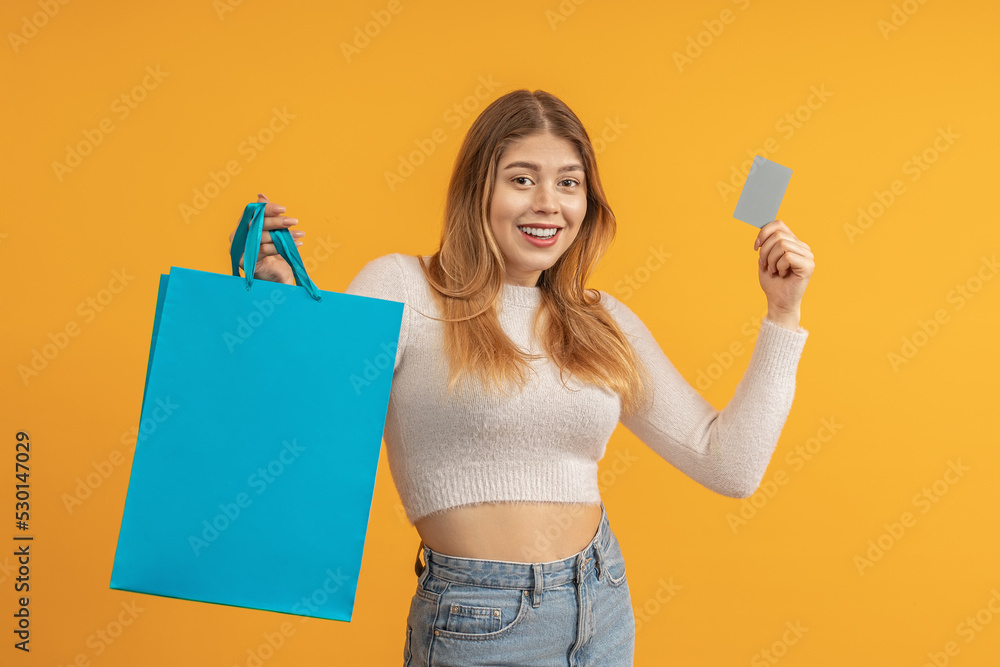 smiling young woman shows a plastic card and holds a paper bag in her hand, stands against a colored background smiling young woman shows a plastic card and holds a paper bag in her hand, stands against a colored background