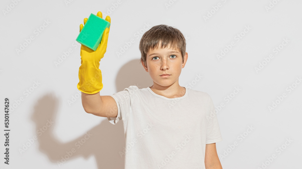 A teenager stands against a white wall, rubber gloves on his hands, a washing sponge in his hand, looks into the camera. Cleaning concept. A teenager stands against a white wall, rubber gloves on his hands, a washing sponge in his hand, looks into the camera. Cleaning concept.