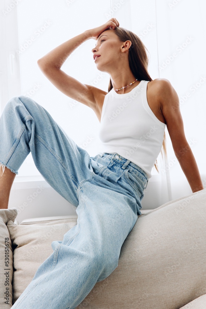A beautiful woman poses on the couch at home in her home clothes in a white tank top and blue jeans