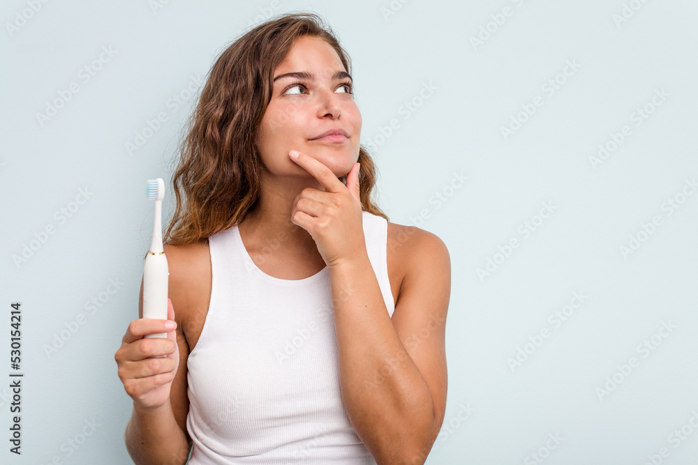 Young caucasian woman holding electric toothbrush isolated blue background looking sideways with doubtful and skeptical expression. Young caucasian woman holding electric toothbrush isolated blue background looking sideways with doubtful and skeptical expression.