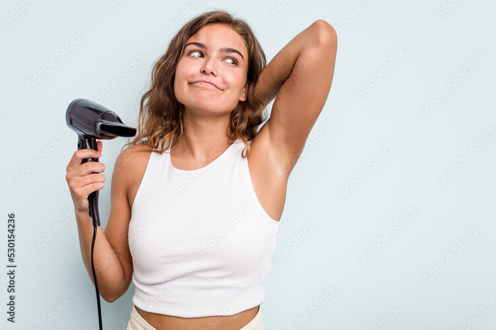 Young caucasian woman holding a hairdryer isolated on blue background touching back of head, thinking and making a choice. Young caucasian woman holding a hairdryer isolated on blue background touching back of head, thinking and making a choice.