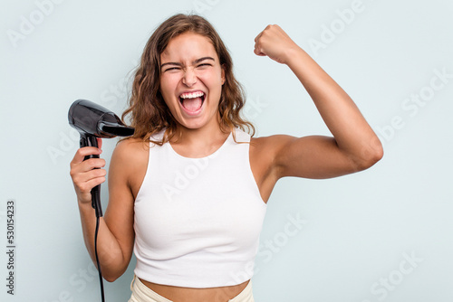 Young caucasian woman holding a hairdryer isolated on blue background raising fist after a victory, winner concept.