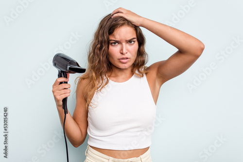 Young caucasian woman holding a hairdryer isolated on blue background being shocked, she has remembered important meeting.