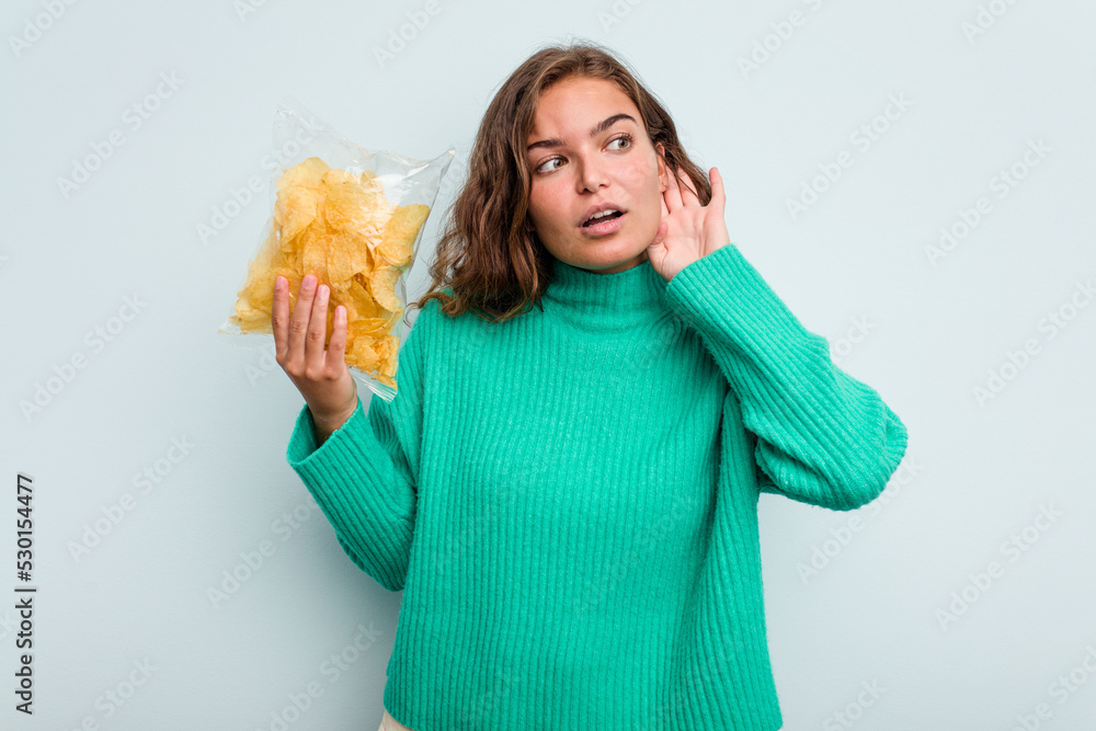 Young caucasian woman holding potato crips isolated on blue background trying to listening a gossip. Young caucasian woman holding potato crips isolated on blue background trying to listening a gossip.